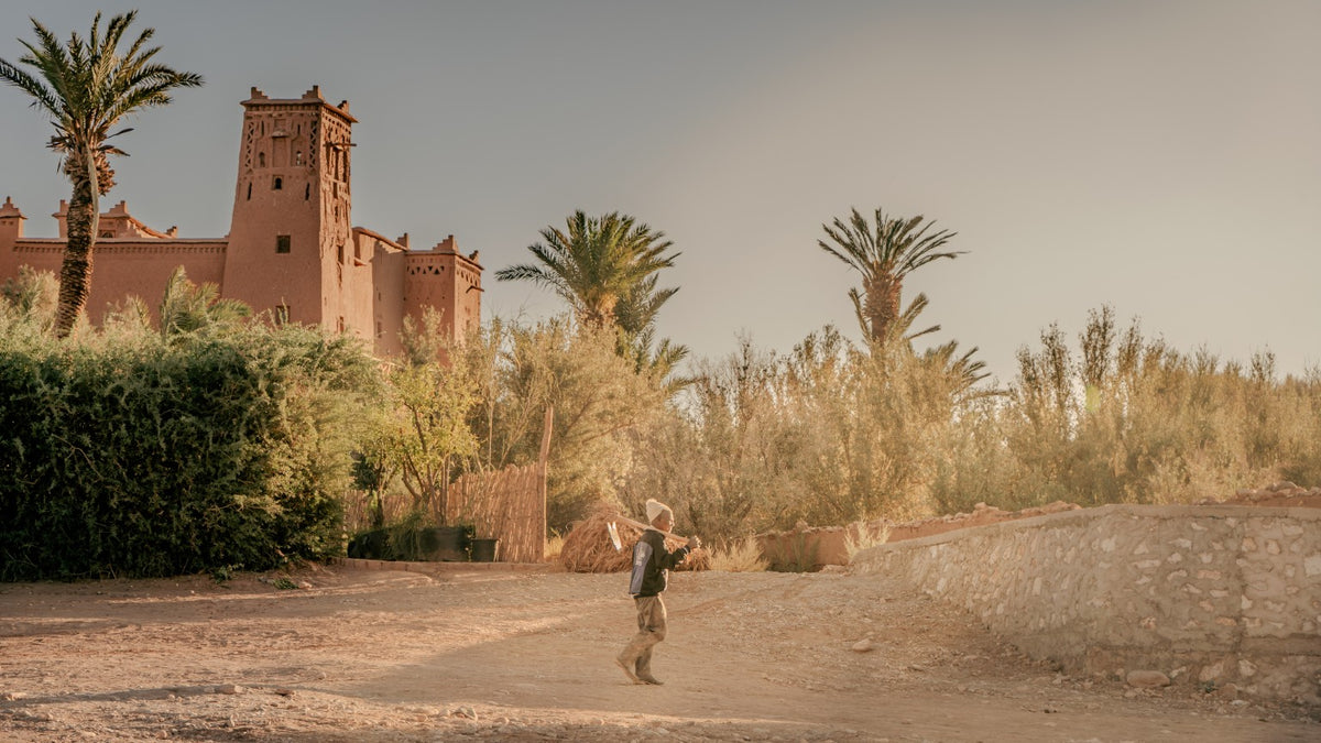 Person walking through the earthy landscape near a Moroccan kasbah and palm trees